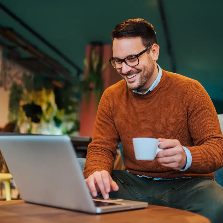 A smiling man holding coffee and using a laptop