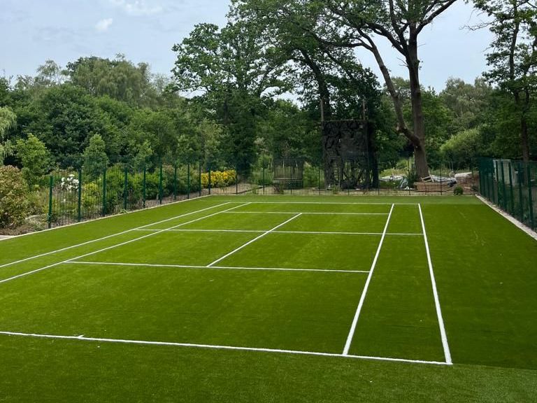 Image shows a green tennis court with white lines with trees around the edges